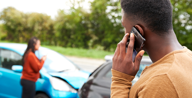 young man on the phone looking at two cars that collided with young woman in the background near the cars; trees behind the road