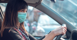 A masked woman sits behind the steering wheel of car, gazing ahead with purpose. 